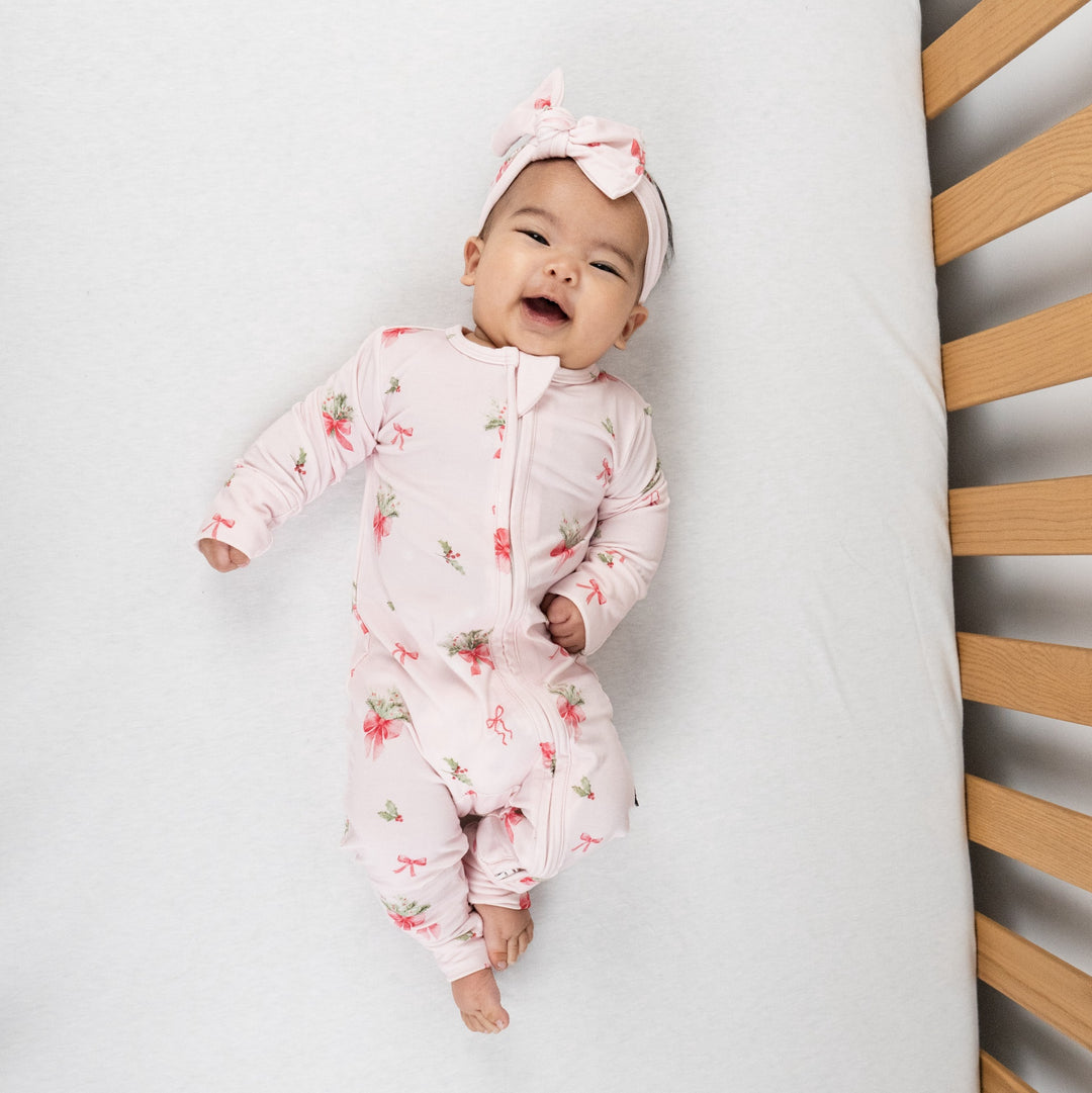 Baby wearing a bamboo footless sleeper pink bows with a pink bow headband, smiling in a crib.