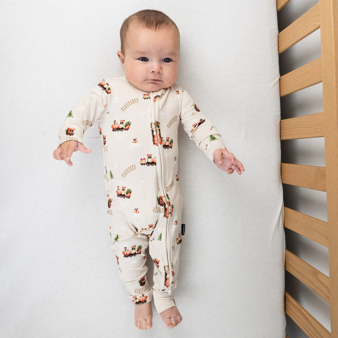 Baby wearing holiday bamboo footless sleeper with a Christmas train print in a crib.