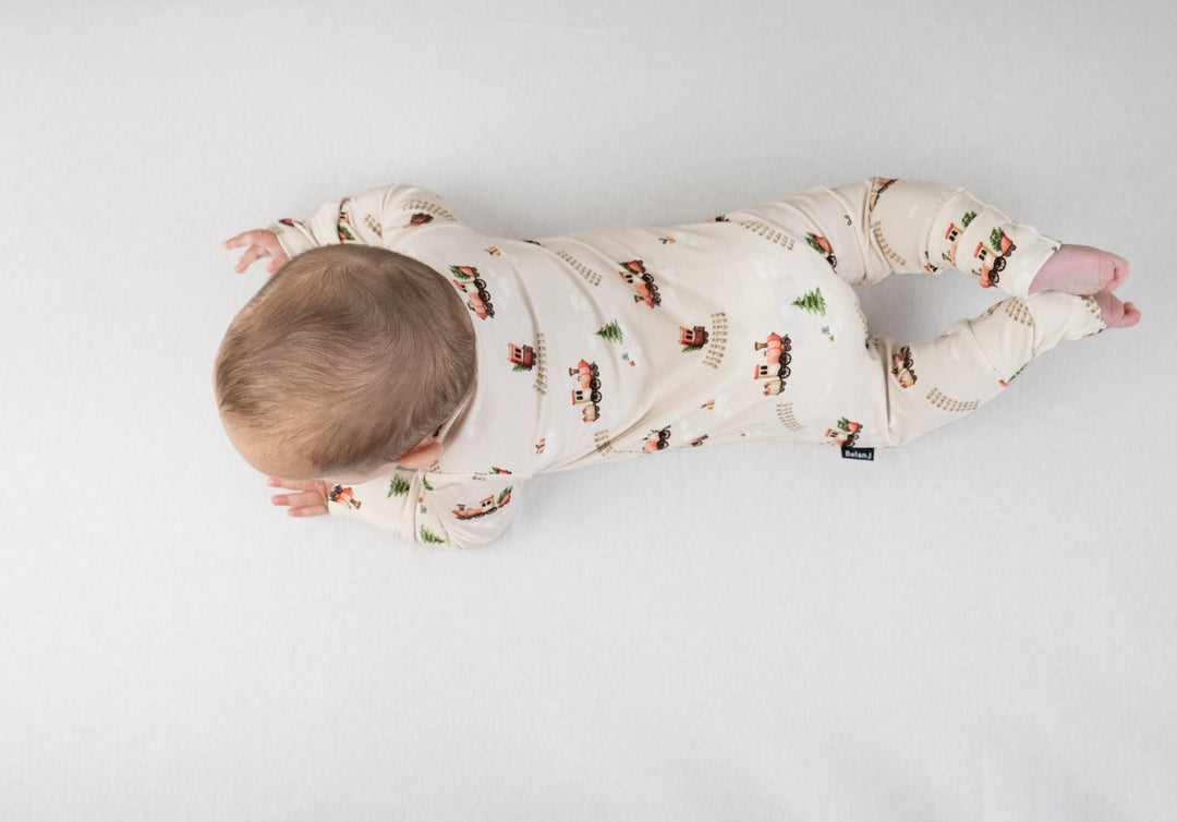 Baby wearing holiday bamboo footless sleeper with Christmas train print, laying on a soft surface.