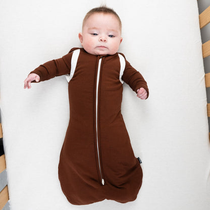 Baby in a cozy 1.0 TOG convertible swaddle sack cocoa, lying on a white mattress.