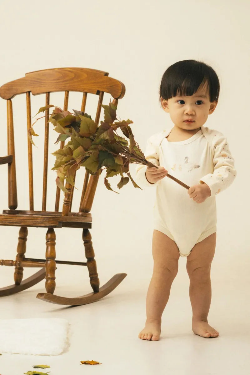Toddler in organic cotton bodysuit holding leaves near a wooden rocking chair, part of the organic cotton baby gift set.